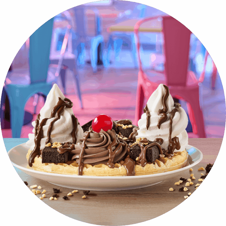 A waffle on a plate, topped with ice cream, brownie pieces, chocolate sauce, and a cherry